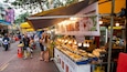 A bustling street food market with various food stalls, people dining, and motorcycles passing by.