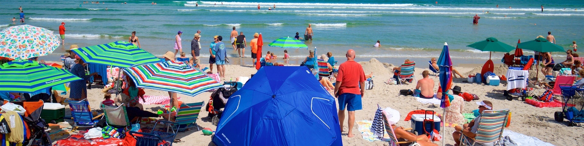 Una playa concurrida con gente tomando el sol, sombrillas y una carpa azul.