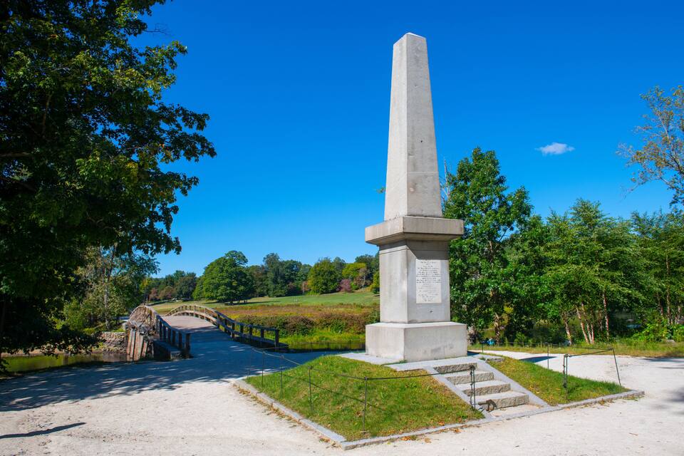A tall obelisk monument in a park with a bridge and greenery.
