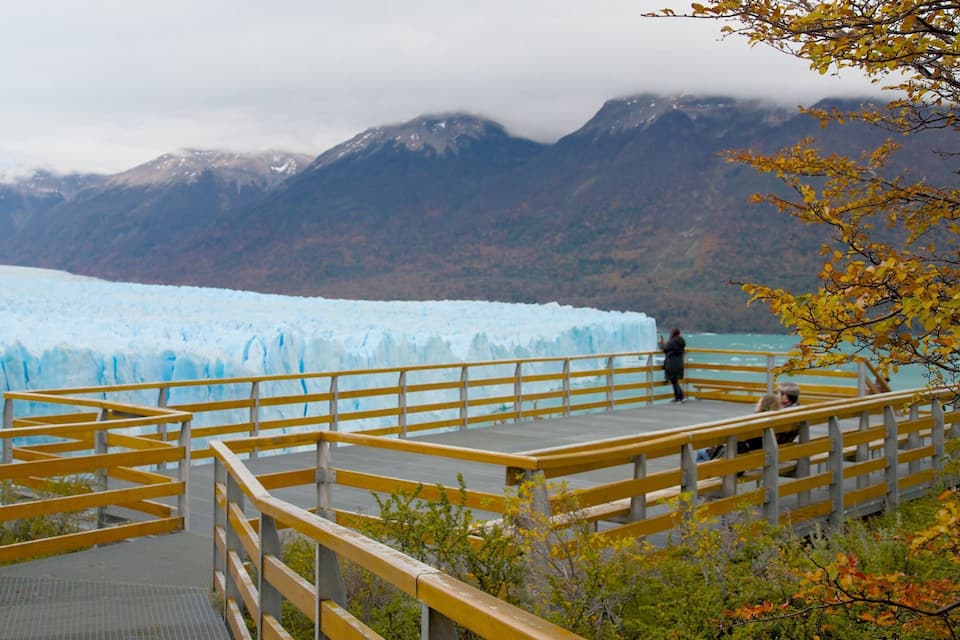 Une plateforme d’observation en bois surplombe un glacier, avec des montagnes en arrière-plan.