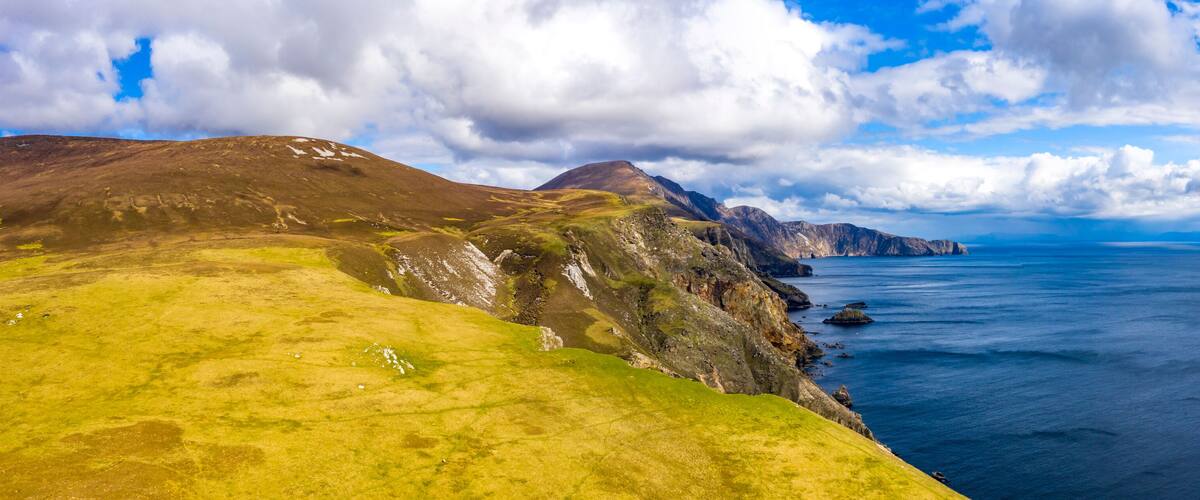 A coastal landscape with grassy hills, rocky cliffs, and a calm sea under a partly cloudy sky.