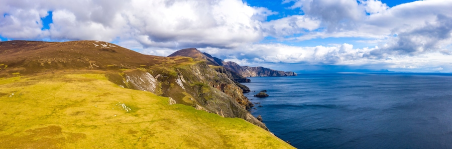 Un paisaje costero con colinas cubiertas de hierba, acantilados rocosos y un mar tranquilo bajo un cielo parcialmente nublado.
