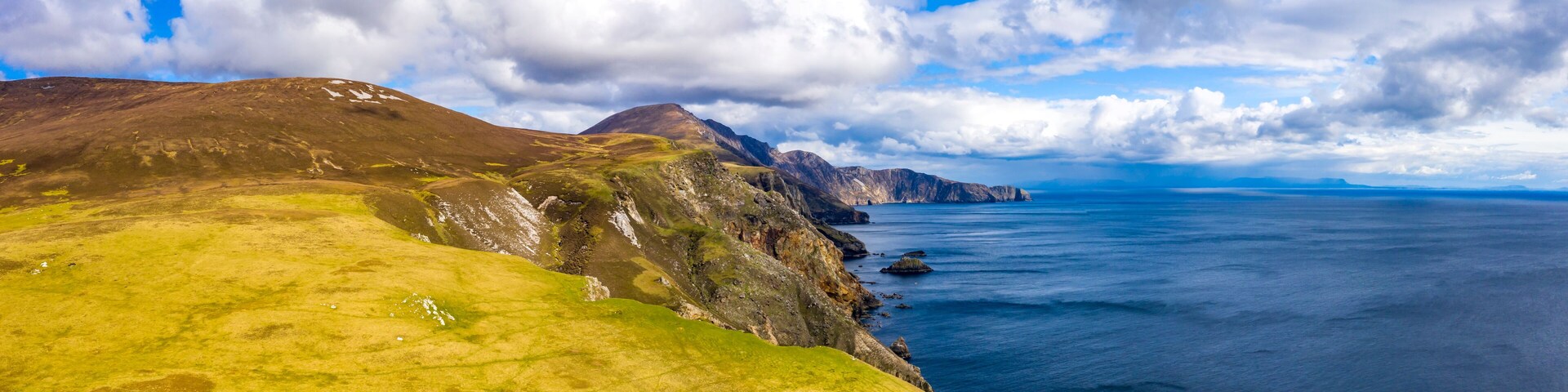 Un paisaje costero con colinas cubiertas de hierba, acantilados rocosos y un mar tranquilo bajo un cielo parcialmente nublado.