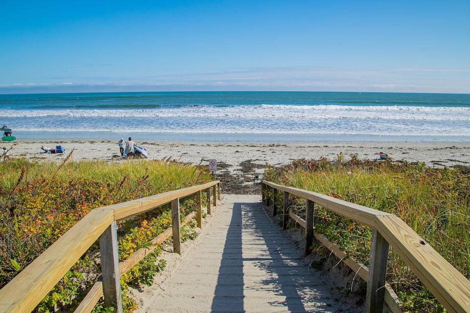 A wooden boardwalk leading to a beach with people and surfboards.