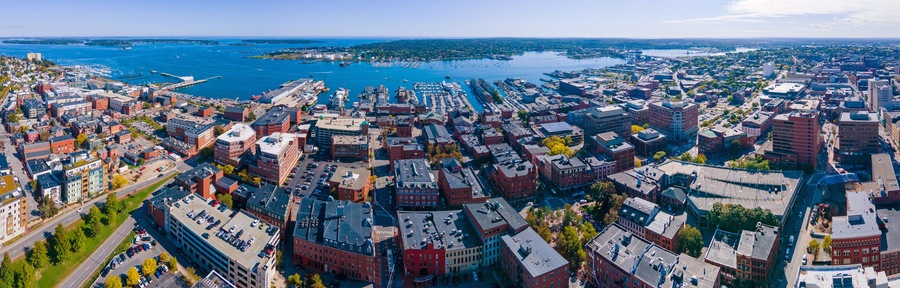 An aerial view of a coastal city with dense urban development, a marina, and a large body of water.