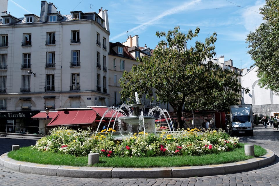 Ein Stadtplatz mit einem Brunnen, umgeben von Blumen, Gebäuden und einem Baum.