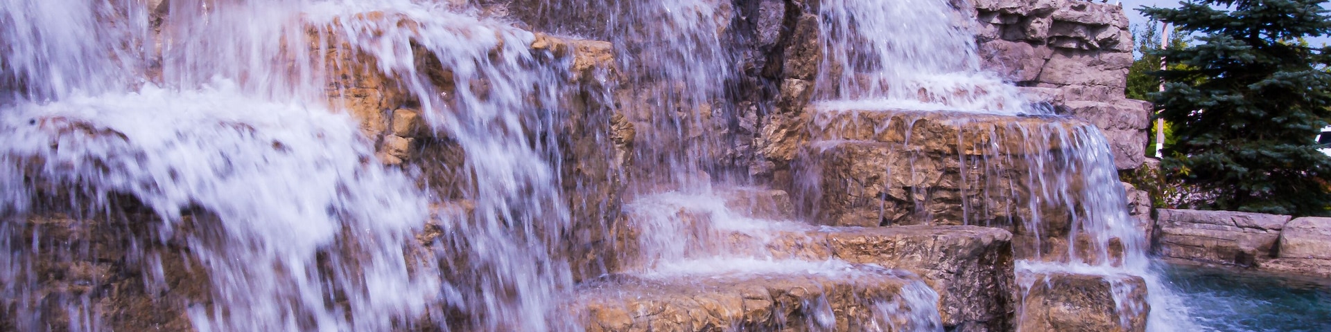 Uma cascata em múltiplos níveis desce sobre um terreno rochoso, com uma árvore de coníferas ao fundo.