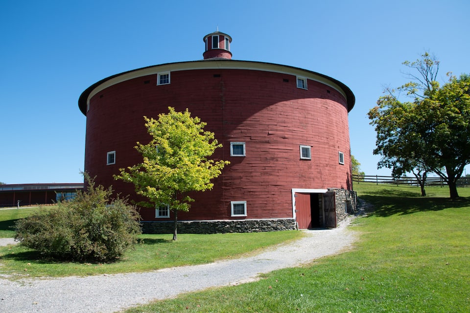 A red cylindrical barn with a stone foundation, a gravel path leading to it, and a tree with yellow leaves nearby.