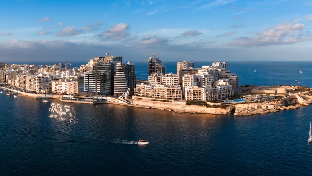 Aerial panorama of Malta at sunset shows St. Julian's and Sliema by Marsamxett Harbour, modern seafront towers, curved promenade, and sailboats crossing deep blue water.