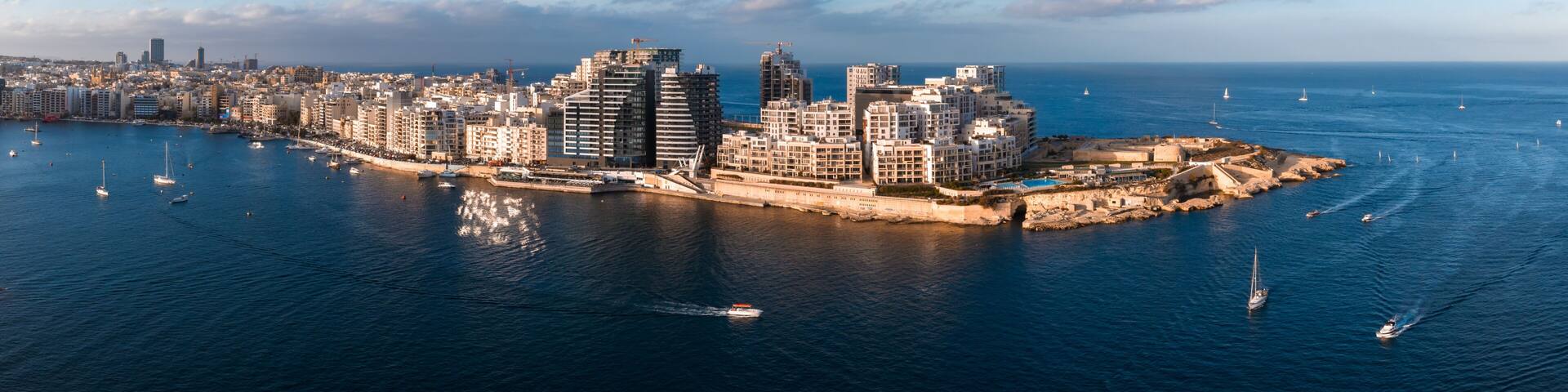 Aerial panorama of Malta at sunset shows St. Julian's and Sliema by Marsamxett Harbour, modern seafront towers, curved promenade, and sailboats crossing deep blue water.