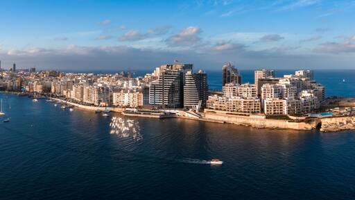 Aerial panorama of Malta at sunset shows St. Julian's and Sliema by Marsamxett Harbour, modern seafront towers, curved promenade, and sailboats crossing deep blue water.
