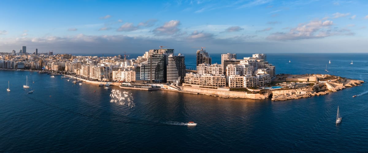 A coastal cityscape with modern buildings, a marina, and sailboats on the water.