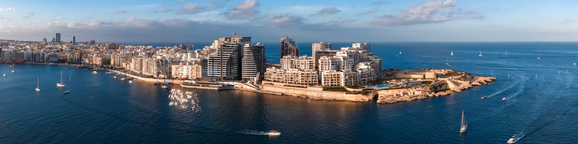 A coastal cityscape with modern buildings, a marina, and sailboats on the water.