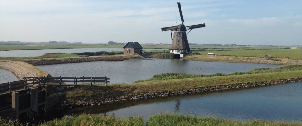 A windmill by a calm pond with a wooden bridge and a small house nearby.