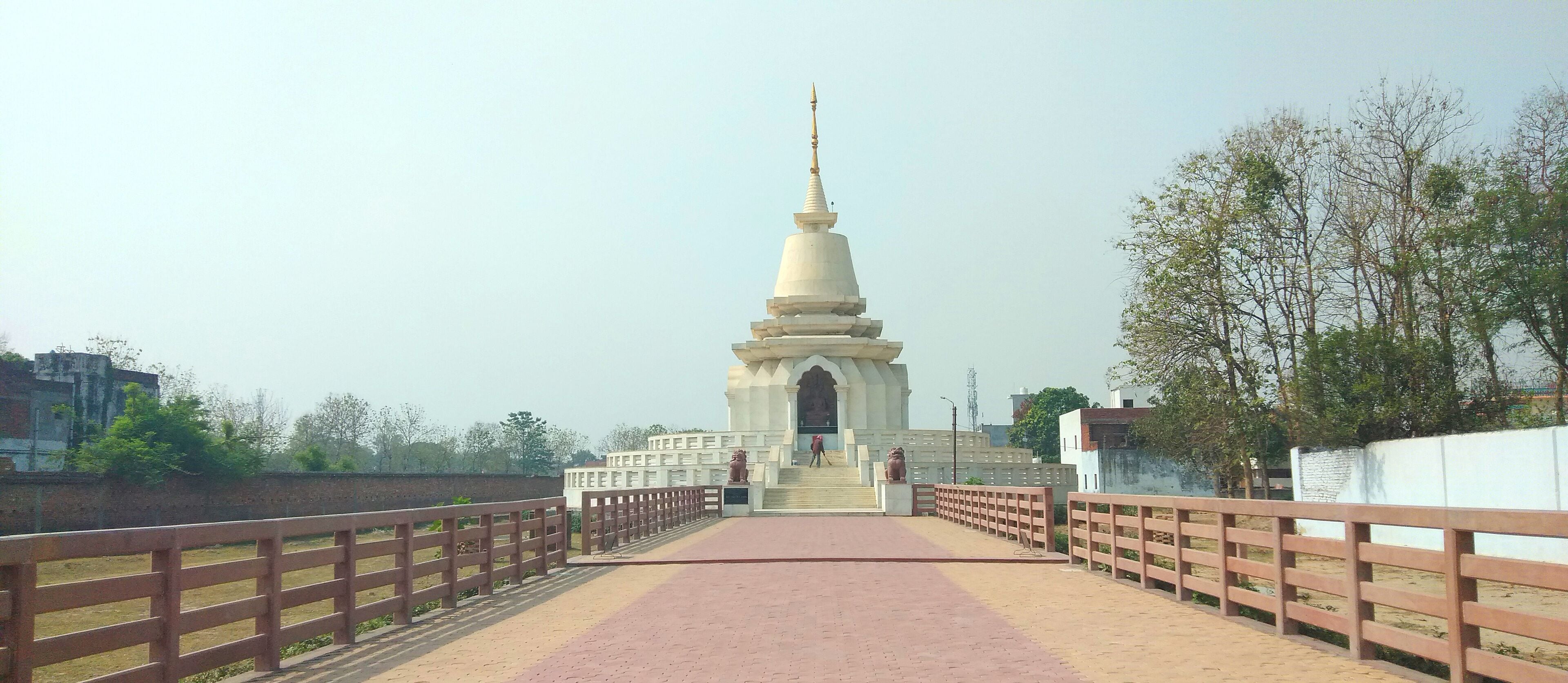 A white stupa with a pointed top, flanked by two large lion statues, on a paved pathway with a wooden railing.