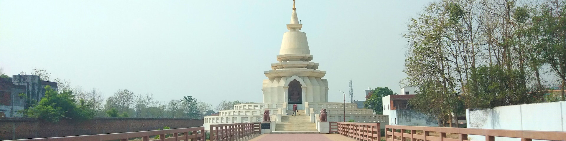 A white stupa with a pointed top, flanked by two large lion statues, on a paved pathway with a wooden railing.