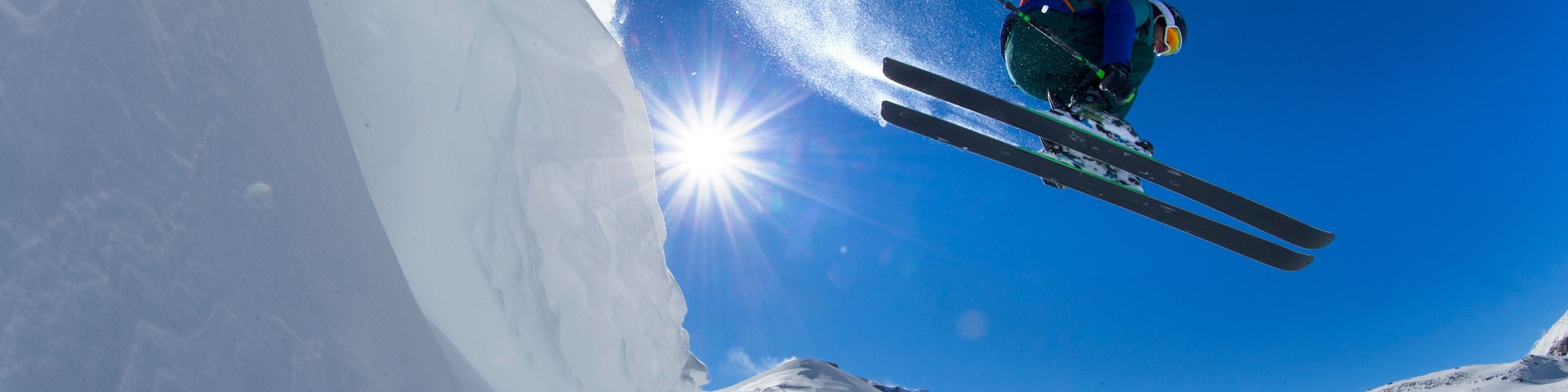 A skier in mid-air performing a jump over a snow-covered slope with the sun shining brightly in the background.