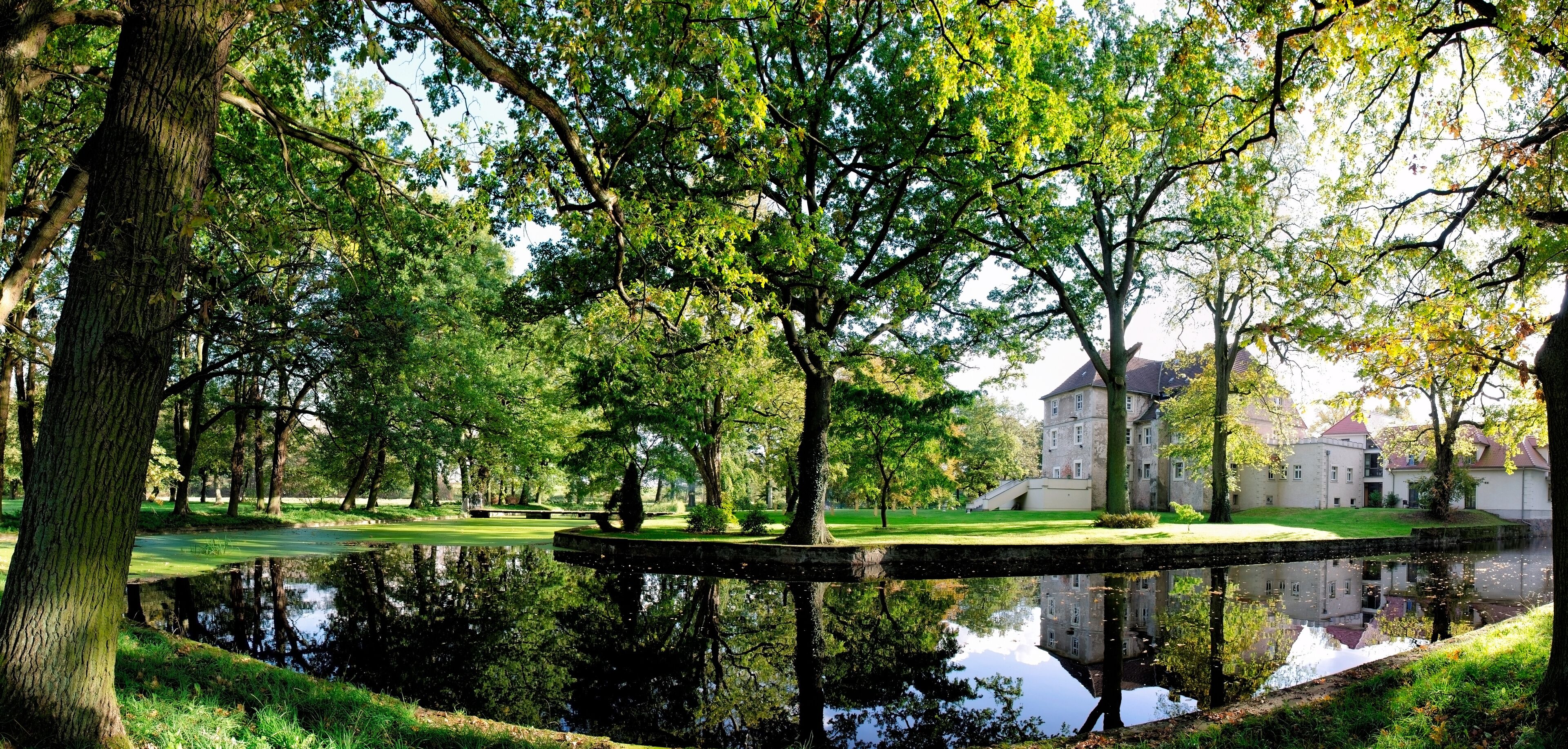 A serene park with a pond, lush trees, and a historic building in the background.