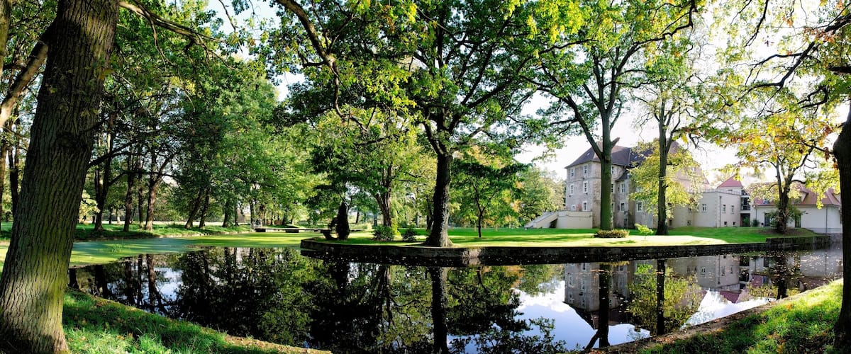A serene park with a pond, lush trees, and a historic building in the background.