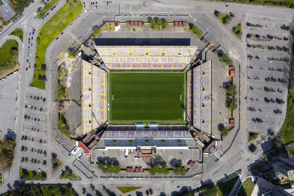 An aerial view of a stadium with a green field, seating areas, and surrounding roads.