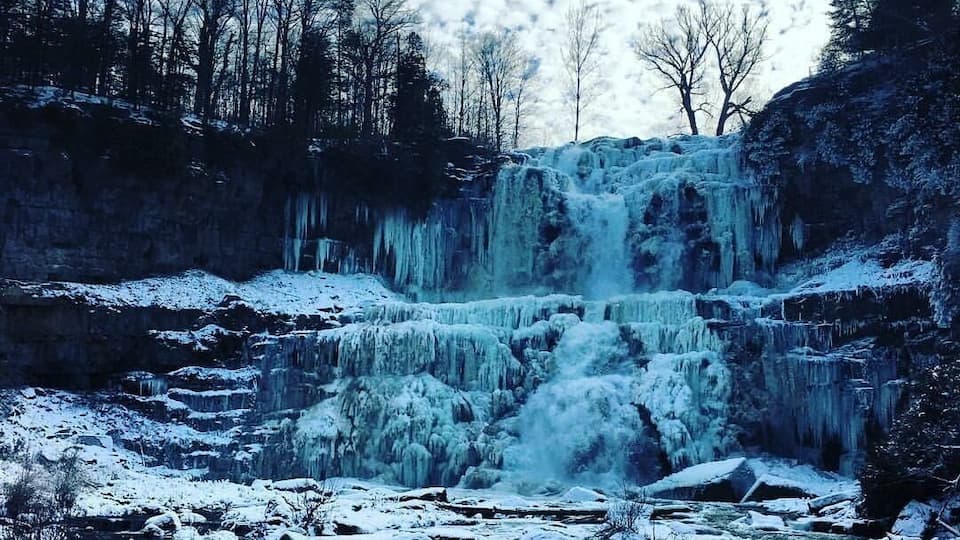 A frozen waterfall in a snowy landscape with bare trees and a river below.