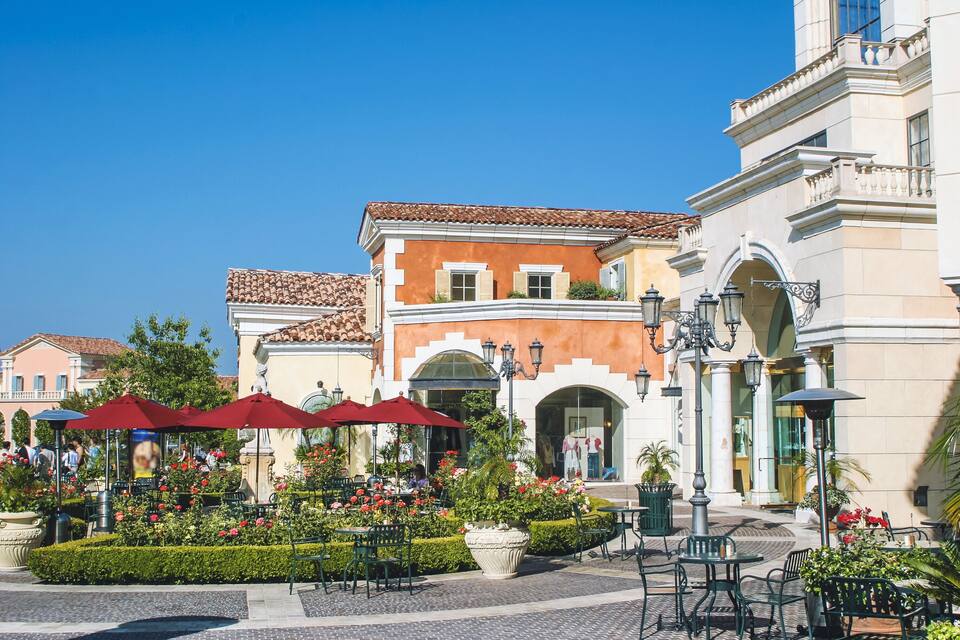 A sunny outdoor cafe with red umbrellas, neatly trimmed hedges, and classic architecture.