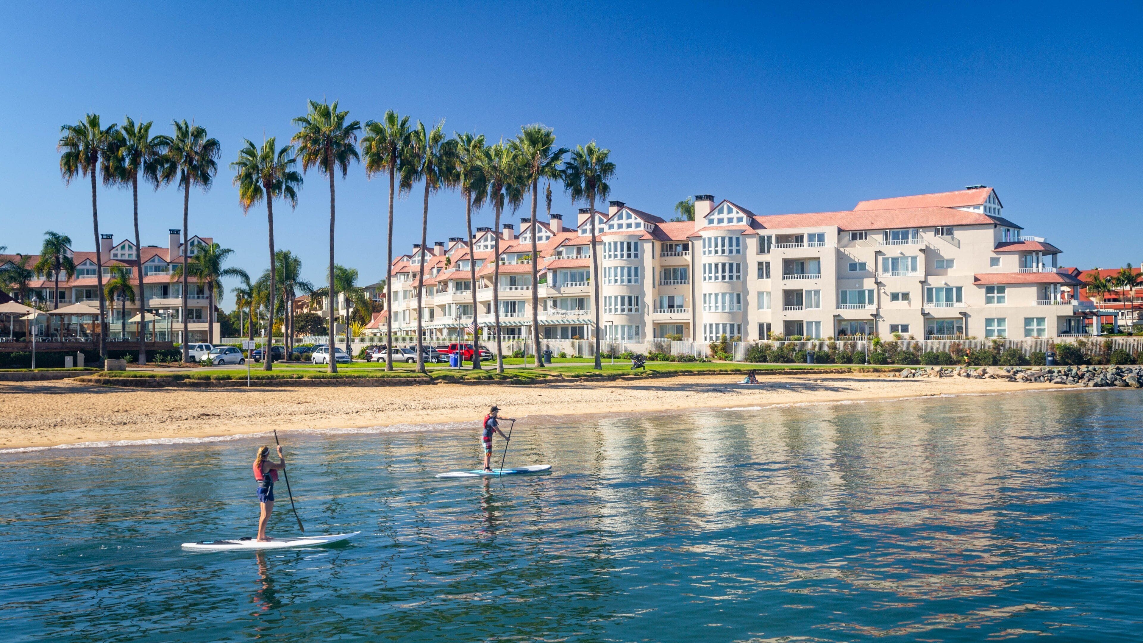 A beach with paddleboarders, palm trees, and a hotel in the background.