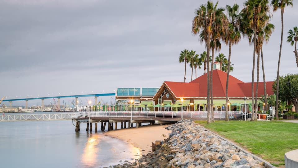 A waterfront restaurant with a red roof, palm trees, and a blue bridge in the background.