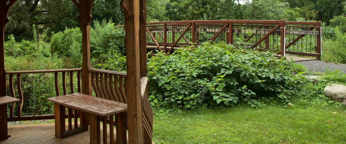 A wooden gazebo with benches, a wooden bridge, and lush greenery.
