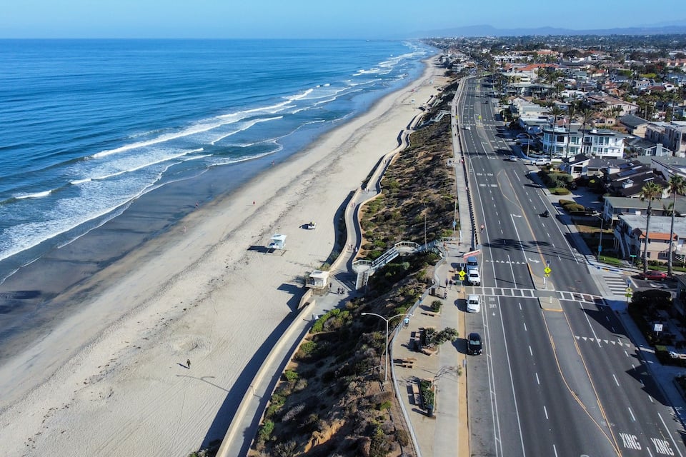 A coastal road with a beach on one side and residential buildings on the other.