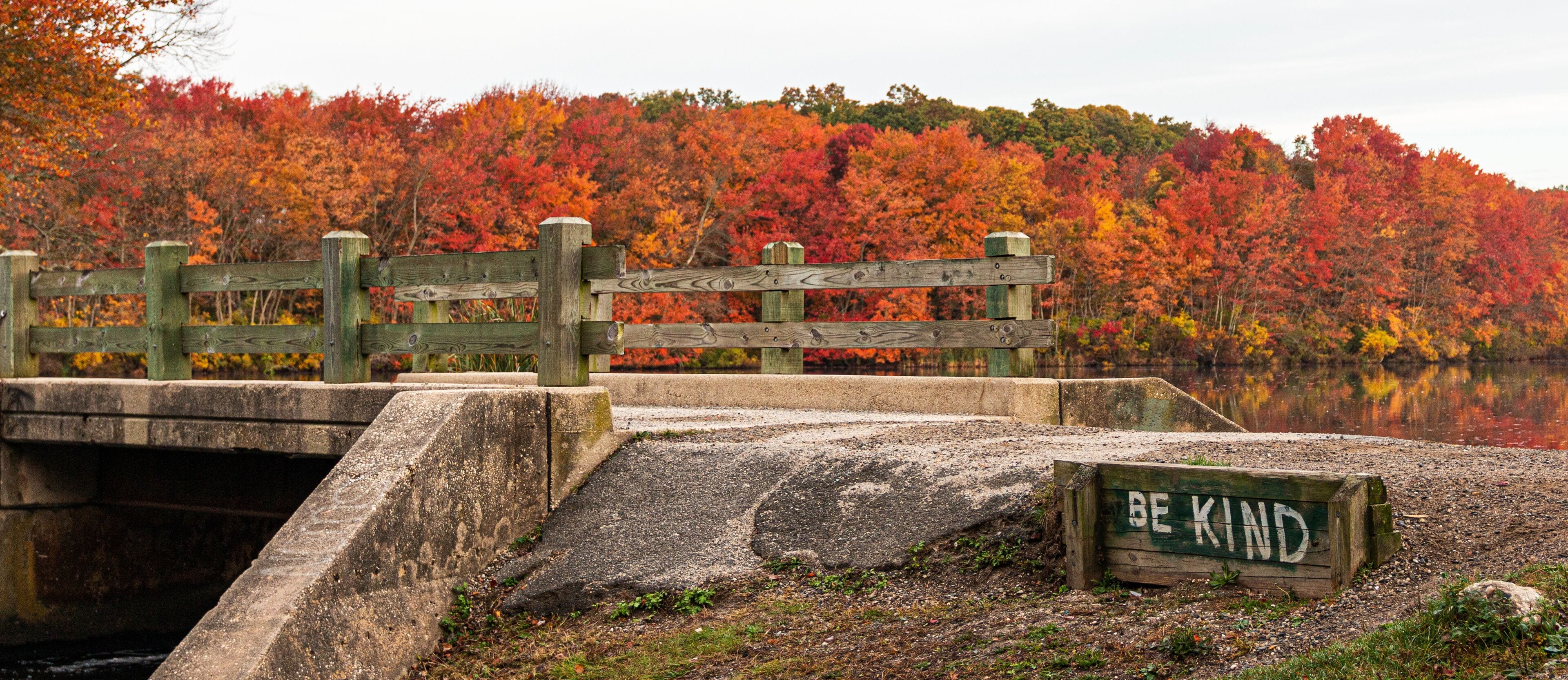 Be Kind is written next to bridge over waterall at Southards Pond with Autumn coloredl leaves in background