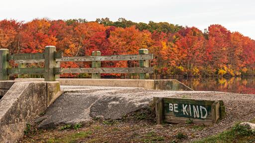 Be Kind is written next to bridge over waterall at Southards Pond with Autumn coloredl leaves in background