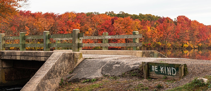 Be Kind is written next to bridge over waterall at Southards Pond with Autumn coloredl leaves in background