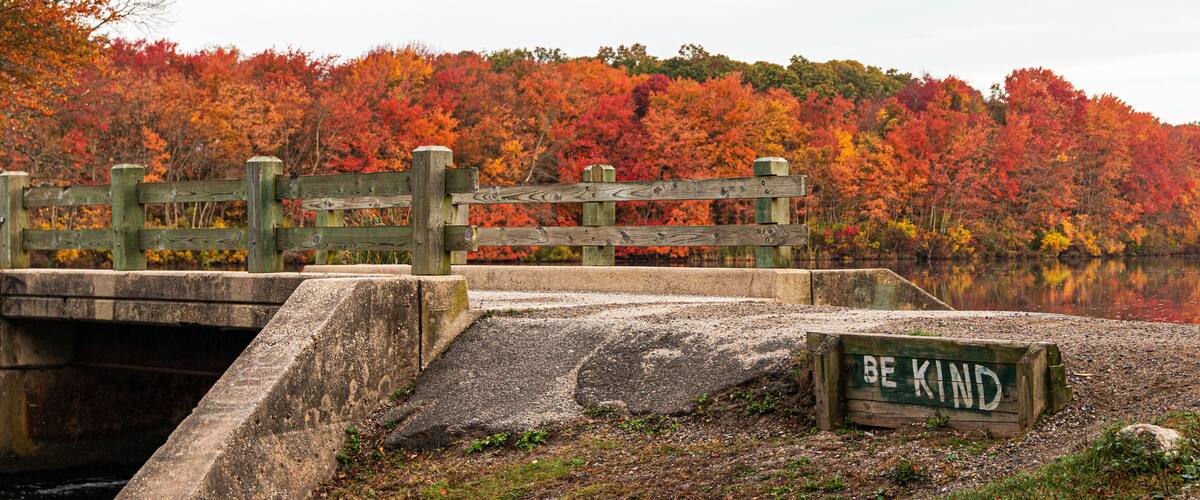A wooden bridge over a body of water with a sign that reads "BE KIND" and a forest with autumn foliage in the background.