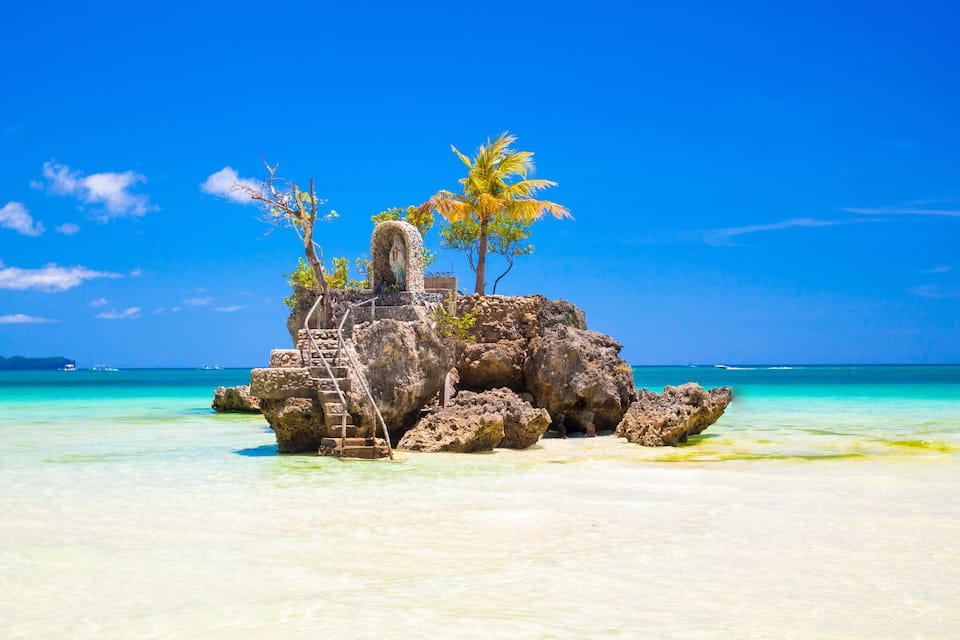 A small rocky island with a statue, palm trees, and clear turquoise waters.