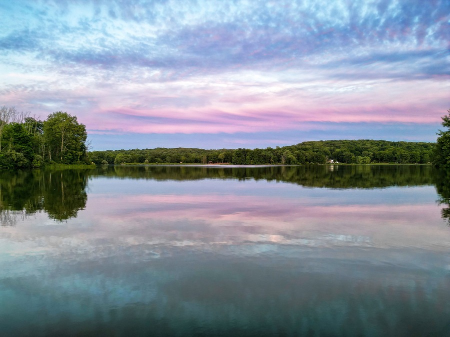 A calm lake with a forested shoreline and a pink and blue sky.