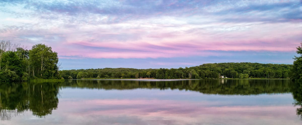 A calm lake with a forested shoreline and a pink and blue sky.