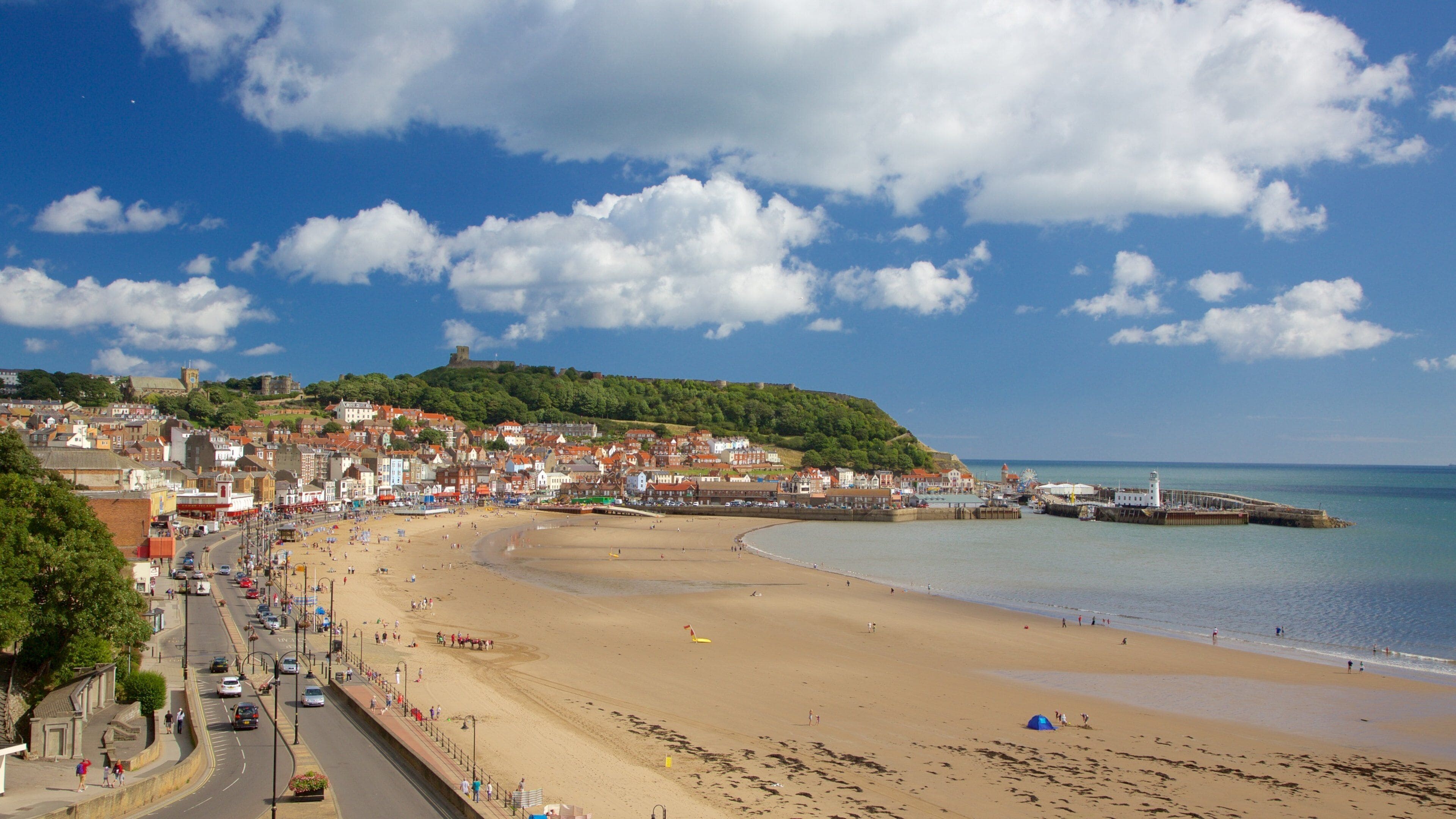 A coastal town with a sandy beach, a historic castle on a hill, and a calm sea.