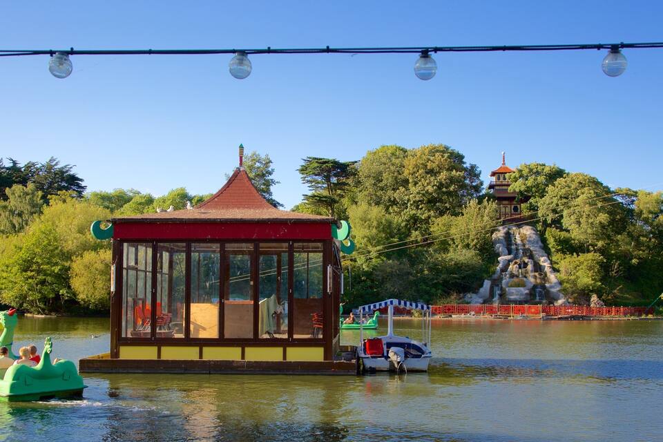 Un pabellón con techo de cristal junto a un lago, un bote verde y una estructura similar a una pagoda al fondo.