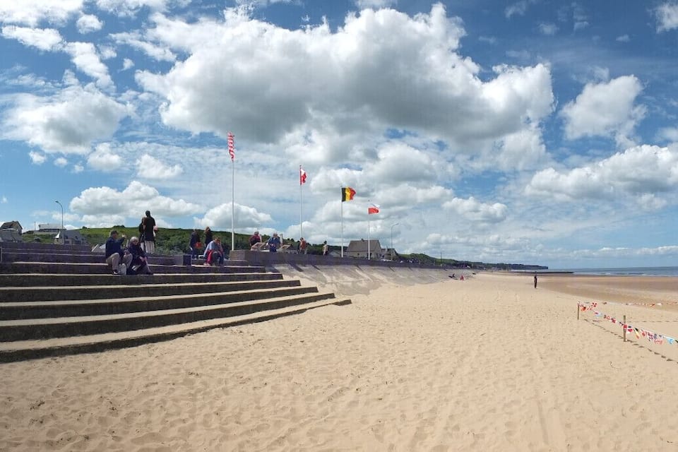 A beach with steps leading down to the shore, a large concrete monument, and a metal sculpture. Flags are flying in the background.