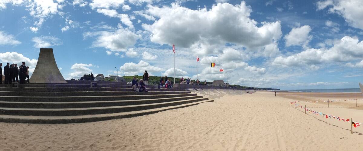 A beach with steps leading down to the shore, a large concrete monument, and a metal sculpture. Flags are flying in the background.