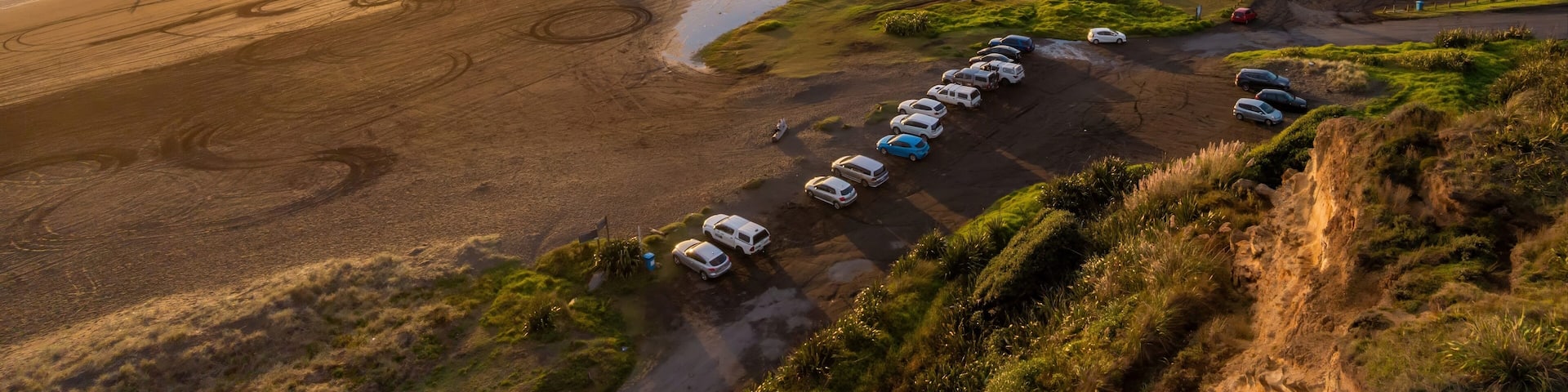Aerial view of Karioitahi Beach, Waiuku, Auckland, New Zealand, at sunset. People are enjoying the beach, some are walking, and others are paragliding. Cars are parked near the beach.