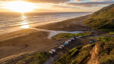 A coastal road with parked cars, a beach, and a sunset.