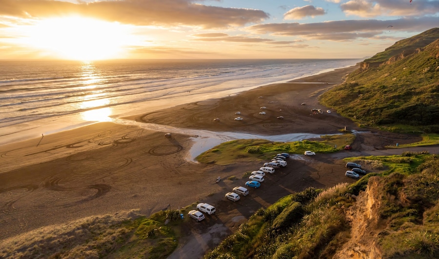 Aerial view of Karioitahi Beach, Waiuku, Auckland, New Zealand, at sunset. People are enjoying the beach, some are walking, and others are paragliding. Cars are parked near the beach.