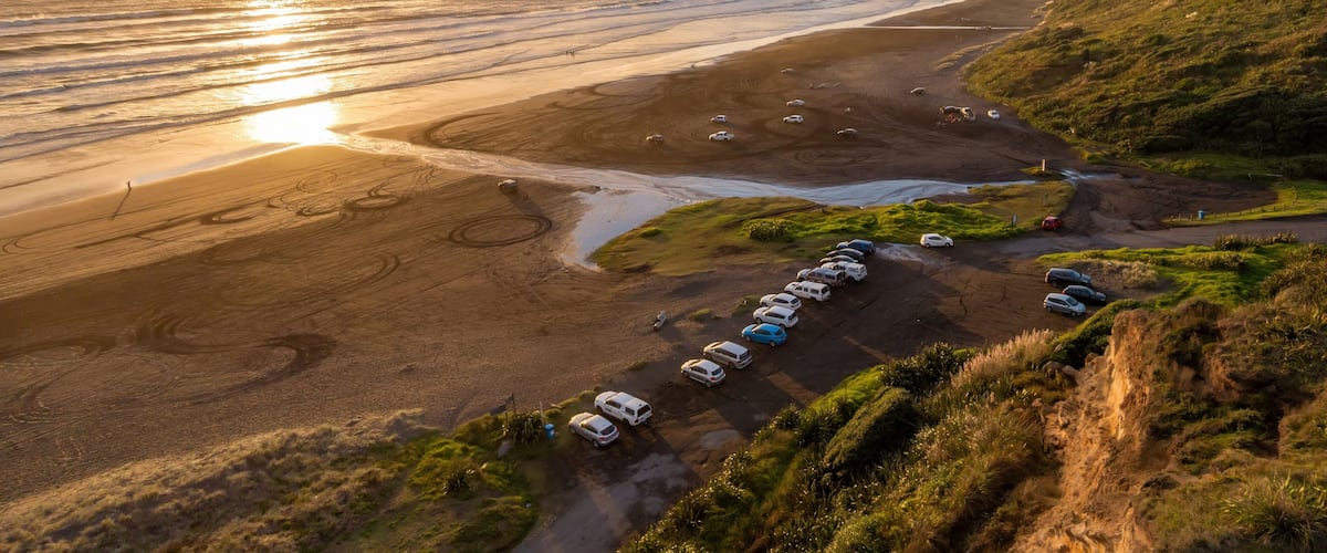 A coastal road with parked cars, a beach, and a sunset.
