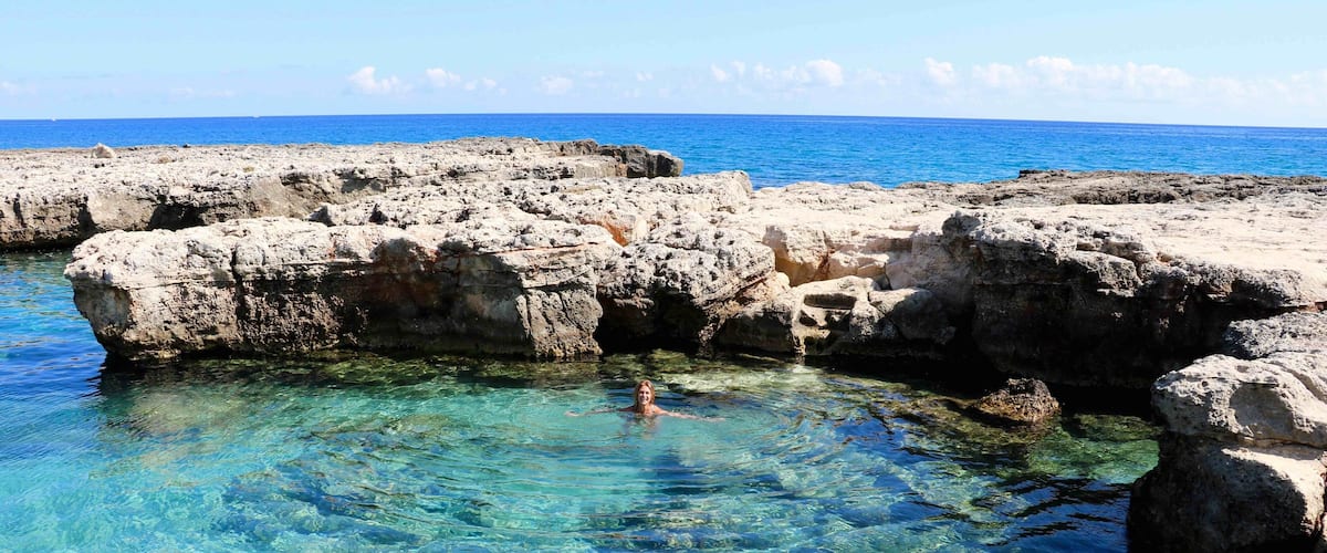 A clear blue sea with rocky formations and a swimmer in the water.