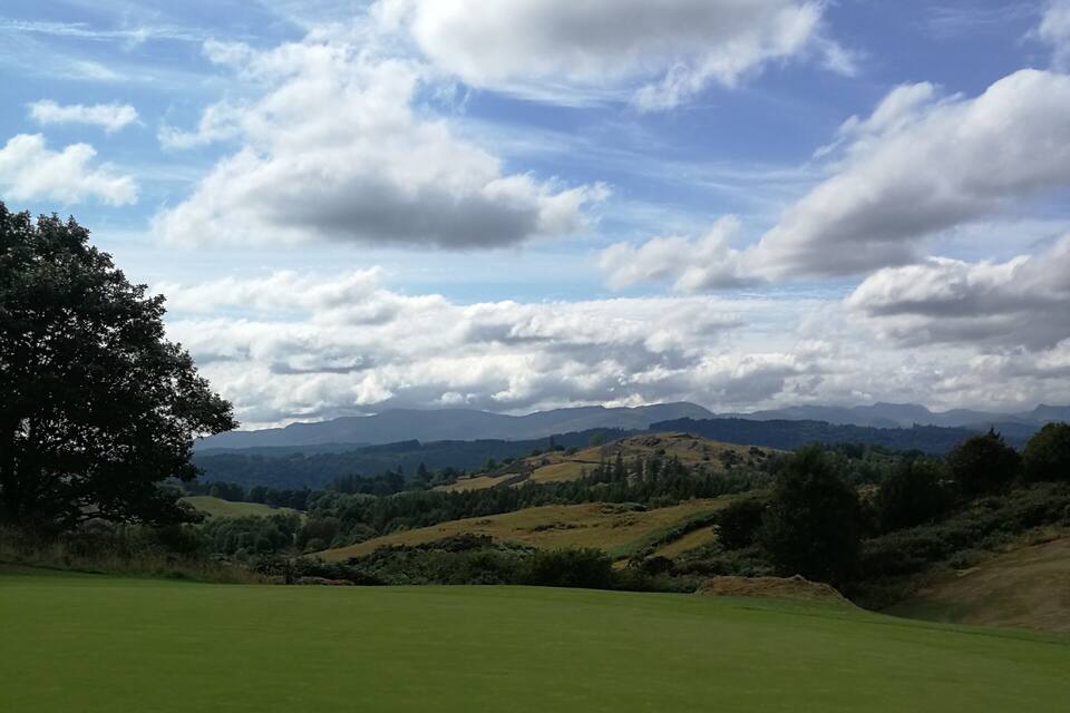 A golf course with a clear sky, rolling hills, and a large tree in the foreground.