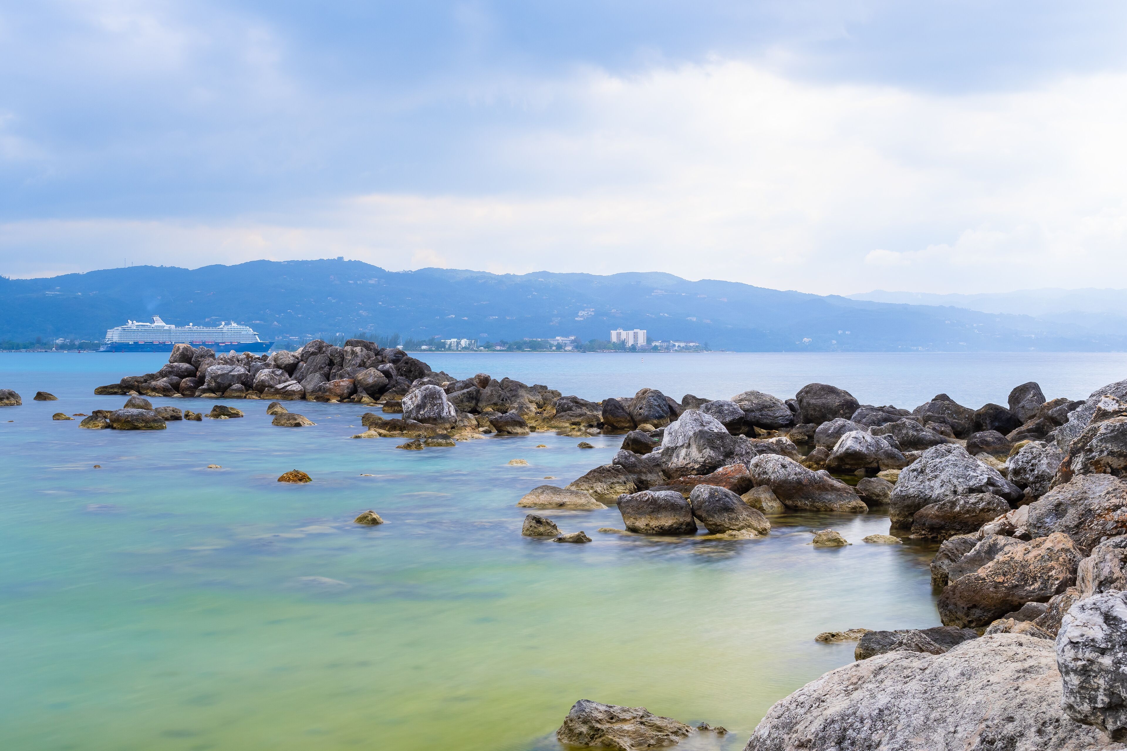 Mein Schiff 6 Cruise Ship docked beyond large ocean rocks in Montego Bay, Jamaica. Mountains in background landscape.