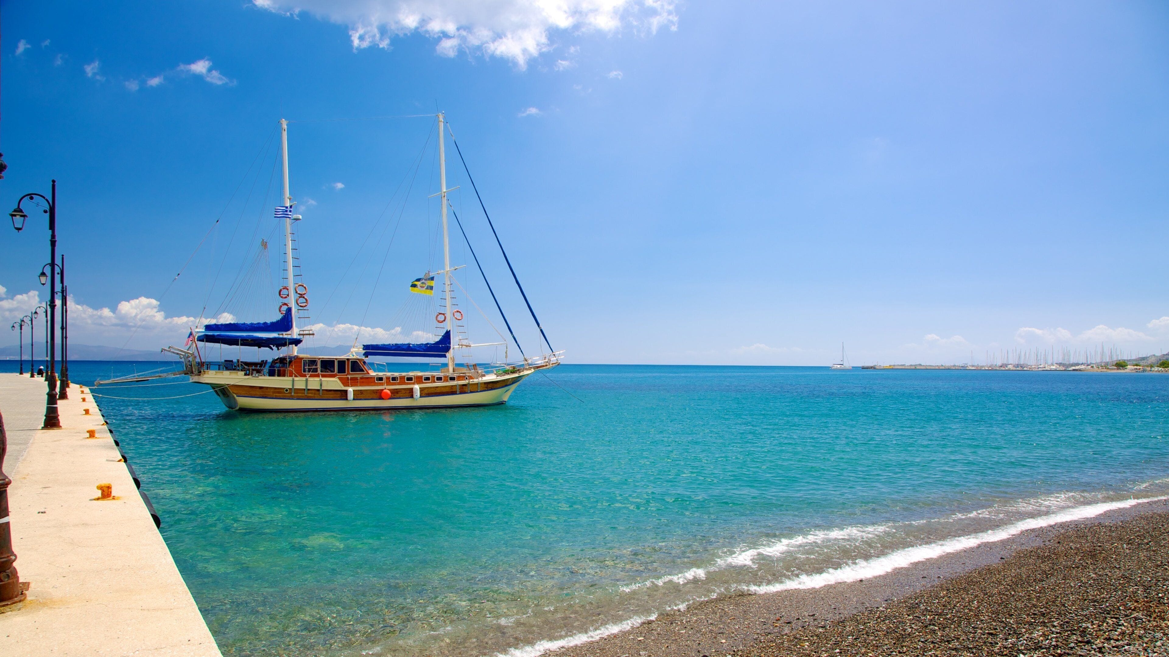 Un barco de madera con banderas atracado en un muelle, agua turquesa cristalina y una playa de guijarros.