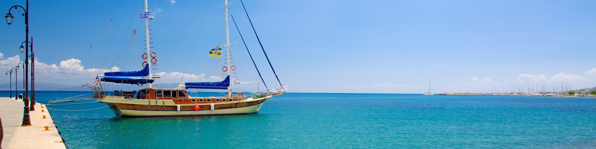 Un barco de madera con banderas atracado en un muelle, agua turquesa cristalina y una playa de guijarros.
